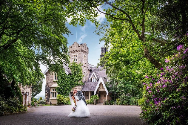 A bride and groom kissing in front of a large manor house covered in ivy