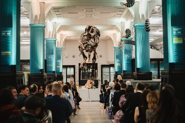 A bride and groom sitting at a table with their friends and family watching. There is a dinosaur skeleton above the couple