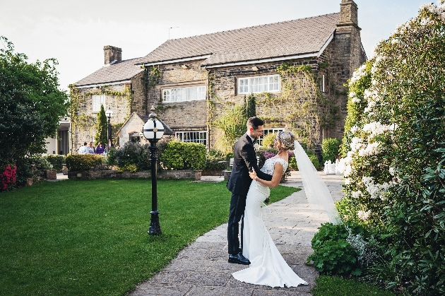 A bride and groom embracing in front of a manor house with grass at the front and flowers