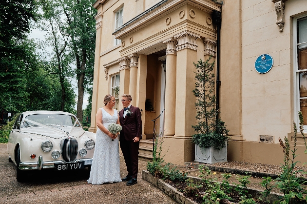 Two people dressed in a dress and suit standing outside of a building with a car in the background